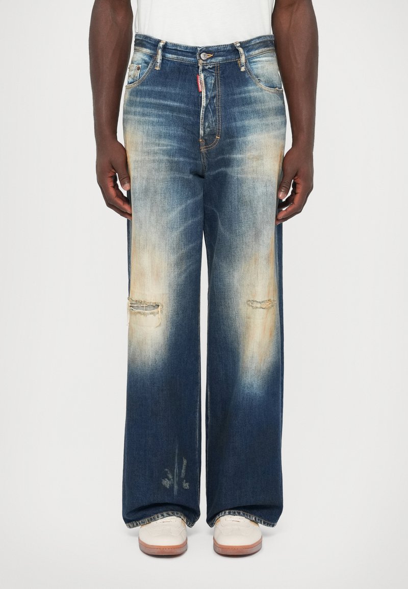 Persona che indossa jeans a gamba larga di un blu sbiadito con buchi strappati sulle ginocchia e sneakers bianche su uno sfondo neutro.
