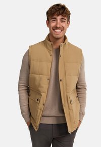Gilet matelassé beige avec un col haut, des boutons-pression, deux poches avant et une texture matelassée. Porté par-dessus un pull gris clair.