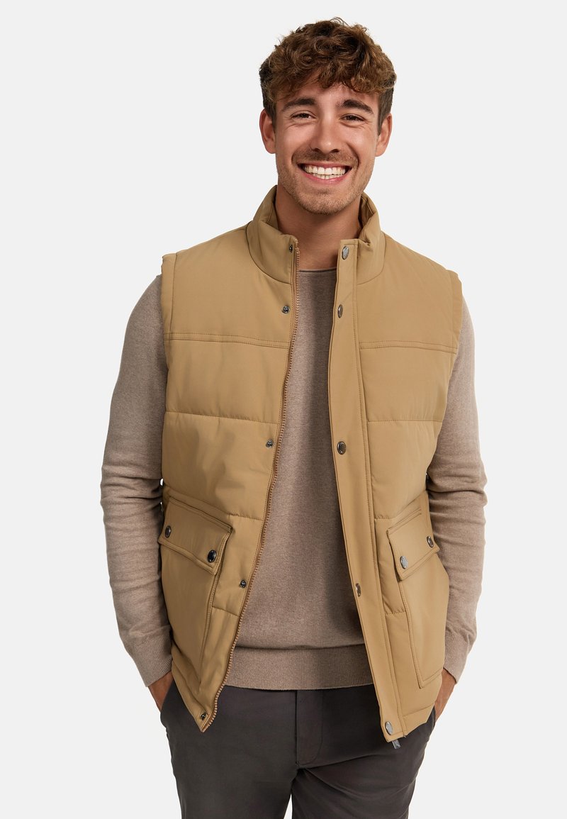 Gilet matelassé beige avec un col haut, des boutons-pression, deux poches avant et une texture matelassée. Porté par-dessus un pull gris clair.