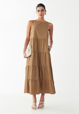 Femme portant une robe longue marron à volants sans manches, tenant une pochette blanche texturée, avec des boucles d'oreilles créoles dorées et des talons blancs à lanières, debout sur un fond blanc.