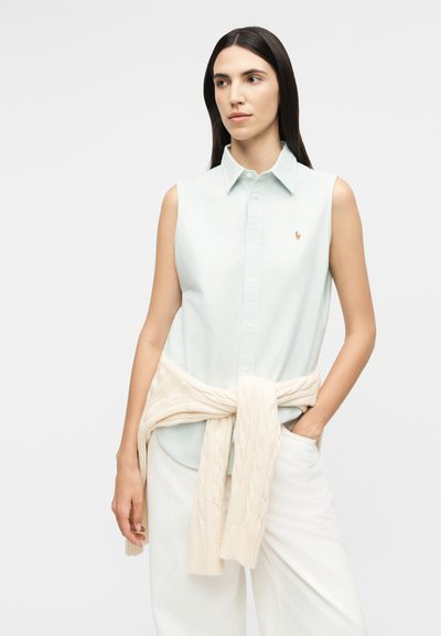 Femme aux longs cheveux foncés portant une chemise sans manches bleu clair boutonnée, un pantalon blanc et un pull en maille torsadée crème noué autour de sa taille.