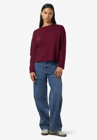 Maglietta a maniche lunghe color mogano con fit rilassato, abbinata a jeans blu a gamba larga. L'outfit presenta un design casual e confortevole con trame morbide.