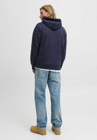 Navy blue hoodie with a drawstring hood, paired with light blue denim jeans and tan footwear, shown from the back against a neutral background.