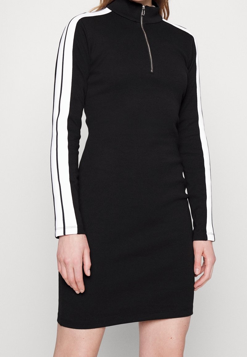 Femme portant une robe noire à manches longues avec des rayures blanches sur les manches et une courte fermeture éclair au col haut.