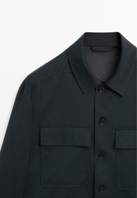 Camicia button-up verde scuro con colletto aperto, due tasche frontali e dettagli di cucitura visibili. Realizzata in tessuto morbido.