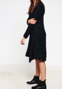 Lång, mörkgrön, fluffig cardigan i texturerat material. Har långa ärmar och en medellång design, kombinerat med en mörk klänning och ankelstövlar.