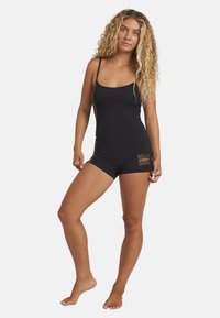 Kobieta z długimi kręconymi blond włosami, ubrana w dopasowany czarny kombinezon bez rękawów z logo "Billabong" na lewym udzie, stoi boso.