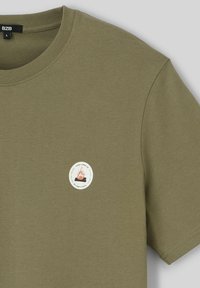 T-shirt en coton vert olive avec un patch logo rond représentant un feu de camp. Encolure ronde basique et manches courtes, texture lisse.