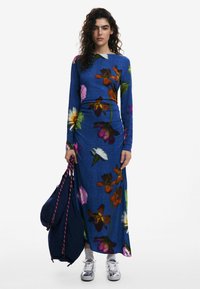 Langärmliges blaues Blumenkleid mit lebhaften Blumen in verschiedenen Rosa-, Orange- und Grüntönen; kombiniert mit einer marineblauen Tasche und silbernen Sneakern.