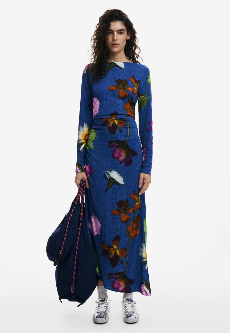 Langärmliges blaues Blumenkleid mit lebhaften Blumen in verschiedenen Rosa-, Orange- und Grüntönen; kombiniert mit einer marineblauen Tasche und silbernen Sneakern.