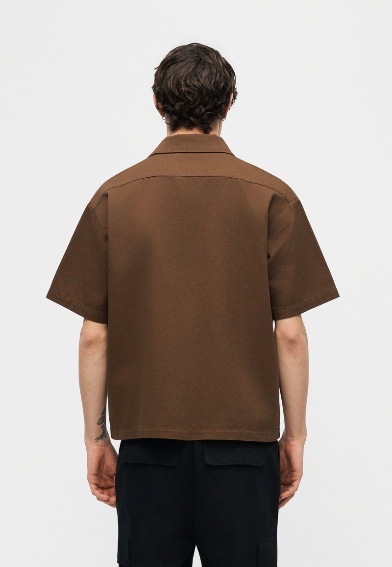 Hombre con cabello corto y rizado, vistiendo una camisa marrón de manga corta y suelta y pantalones negros, de pie de espaldas sobre un fondo liso.