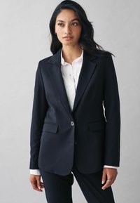 Sort skræddersyet blazer i et glat stof med to frontlommer, enkeltknaplukning og slagklapper, båret over en hvid skjorte.