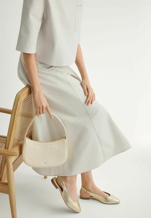 Osoba siedząca na drewnianym krześle, ubrana w jasnobeżową midi spódnicę i top, trzymająca kremową torebkę, na stopach złote baleriny na pasku.
