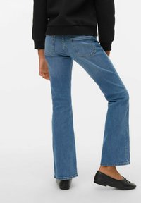 Osoba nosící světle modré džínové kalhoty s rozšířenými nohavicemi, černý svetr a černé baleríny, stojící s jednou nohou mírně pokrčenou.