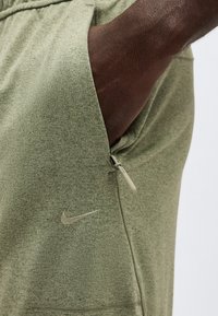 Χέρι που έχει εισαχθεί στην πλαϊνή τσέπη με φερμουάρ σε ελαφρώς πράσινο-ελαιώδες παντελόνι Nike, δείχνοντας την υφή του υφάσματος και την κεντημένη λογότυπο.