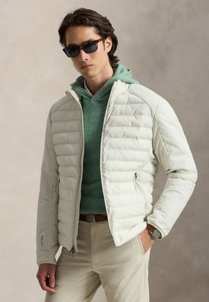 Homme portant des lunettes de soleil noires, une veste matelassée beige clair, un sweat à capuche vert, un pantalon beige et une montre-bracelet, debout avec une main dans la poche.