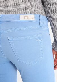 Světlé modré džíny z denimu s hladkou texturou, které mají zadní kapsy a nášivku s logem LTB na pase. Detailní pohled s pleteným svetrem.