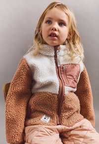 Jachetă din fleece cu un design în blocuri de culoare albă și maro, având un buzunar pe piept cu fermoar și un finisaj moale, texturat.