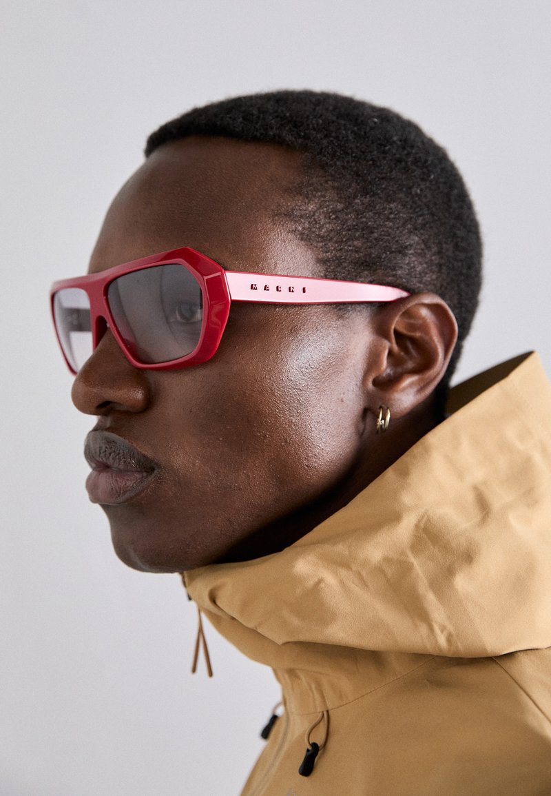 Rote, eckige Sonnenbrille mit einem dicken Rahmen und getönten Gläsern. Präsentiert an einem Model, das eine beigefarbene Steppjacke mit hohem Kragen trägt.