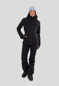 Czarna ocieplana kurtka z zapięciem na zamek i kapturem, zestawiona z czarnymi spodniami narciarskimi. Jasnoniebieska czapka dopełnia zimowy strój.
