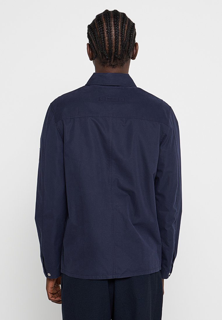 Chaqueta de manga larga azul marino con cuello, de corte recto, puños abotonados y una sutil textura en la tela. Detalles de costura visibles.