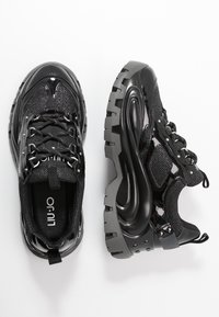 Černé tenisky s lesklým, lakovaným povrchem a sítovanou tkaninou. Masivní podrážka s texturovaným vzorem a kovovými prvky na tkaničkách.