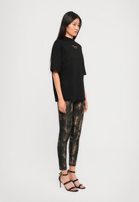 T-shirt nera oversize con testo dorato, abbinata a leggings neri con motivo rame. Sandali alti neri con affini completano l'outfit.