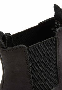 Botín negro con cuero liso y paneles de malla texturada. Forma redondeada con un borde cosido y una lengüeta para facilitar el calzado.