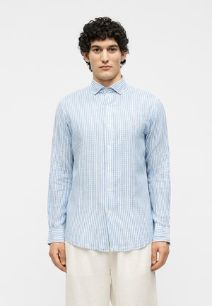 Νέος άνδρας με κοντά σγουρά μαλλιά, που φοράει ένα πουκάμισο με κάθετες μπλε και λευκές ρίγες και κρεμ παντελόνι, στέκεται μπροστά σε λευκό φόντο.