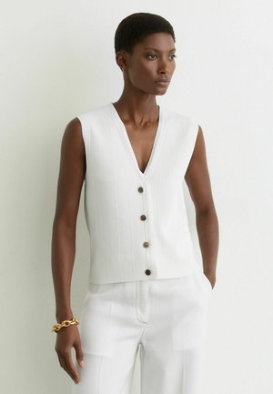 Nainen, jolla on valkoinen hihaton napillinen liivi ja valkoiset housut, seisoo vaaleaa seinää vasten ja vasemmassa ranteessa on kultainen ketjurannekoru.