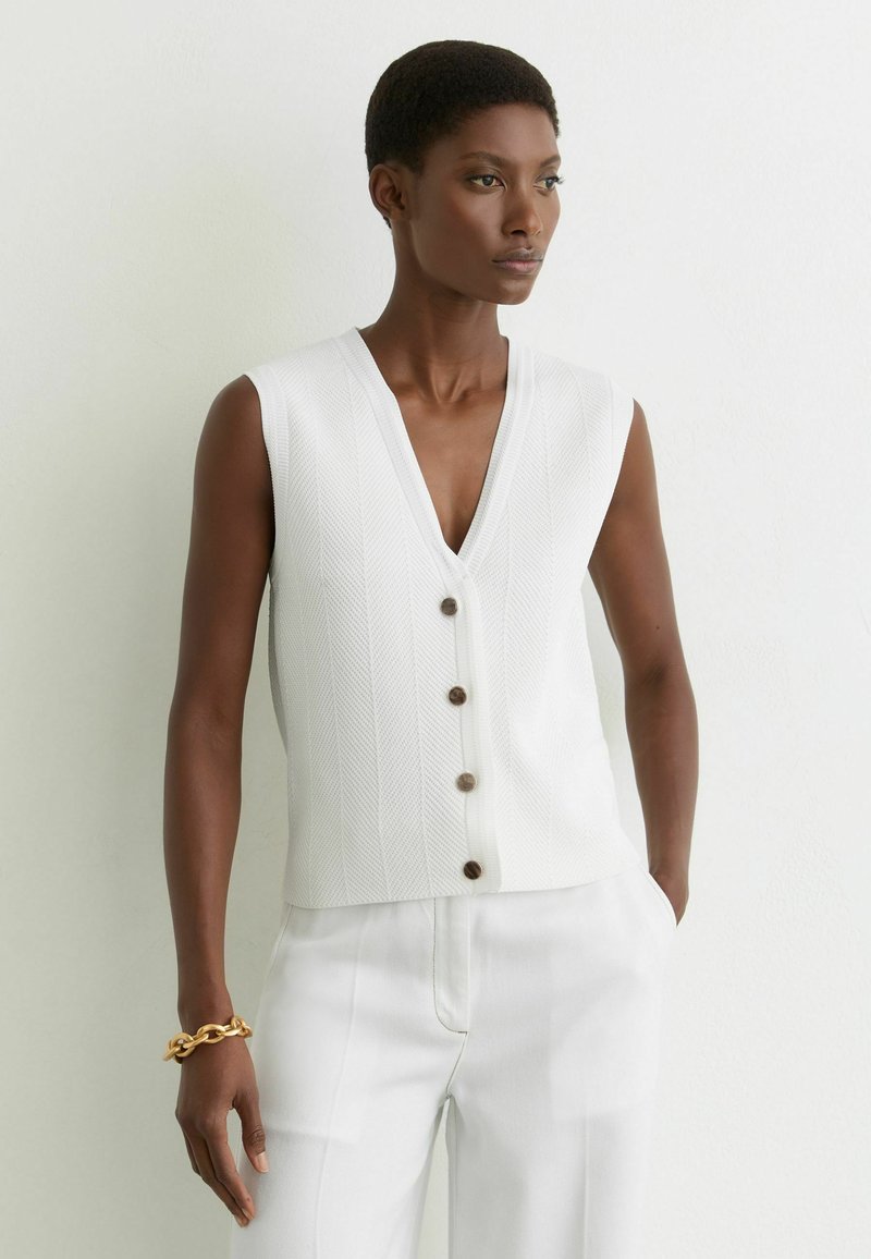 Vrouw met een mouwloze witte blouse met knopen en witte broek, staand tegen een effen lichte muur met een gouden armband aan haar linker pols.