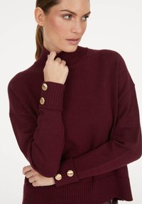 Burgunderfarbener Strickpullover mit hohem Kragen, geripptem Design und goldenen Knopfakzenten an den Bündchen. Lockere Passform mit seitlichen Schlitzen am Saum.
