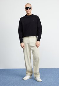 Svart hettegenser med en liten logo, beige avslappede bukser og hvite joggesko, kombinert med store, mørke solbriller. Jevnt stoff og moderne design.