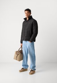 Chaqueta de plumas negra con cuello alto y cremallera. Jeans de mezclilla azul claro, calzado beige y un bolso marrón claro con un acento marrón oscuro.