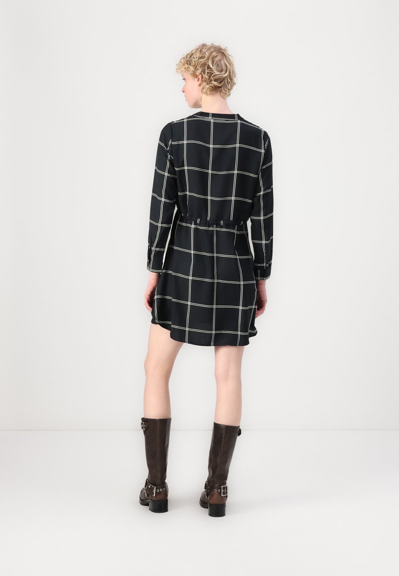 Schwarzes Karokleid mit langen Ärmeln, tailliertem Bund und ausgestelltem Saum, kombiniert mit knielangen braunen Lederstiefeln.