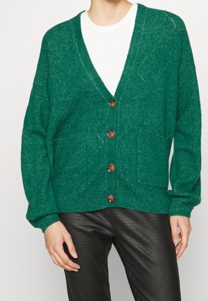 Personne portant un cardigan vert boutonné avec des poches avant, par-dessus une chemise blanche et un pantalon noir texturé, debout devant un fond uni.