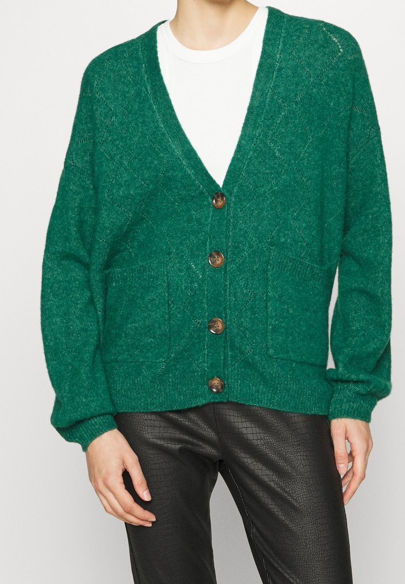 Personne portant un cardigan vert boutonné avec des poches avant, par-dessus une chemise blanche et un pantalon noir texturé, debout devant un fond uni.