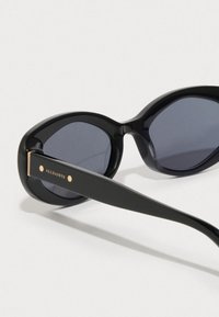 Čierne slnečné okuliare s nadrozmerným oválnym rámom, tmavými sklami a matným povrchom. Obsahujú zlatý akcent na spánku označený "ALLSAINTS."