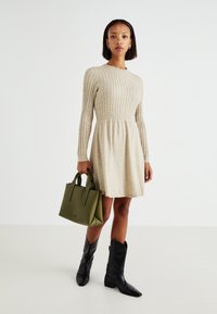 Robe en tricot beige avec une texture côtelée, des manches longues et une jupe évasée. Associée à un sac à main en cuir vert et des bottes noires à bouts pointus.
