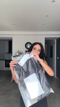 Μαύρο ένδυμα σε διαφανή πλαστική σακούλα, με ετικέτες. Η σακούλα έχει μια λευκή ετικέτα με κείμενο. Ο εσωτερικός χώρος έχει μοντέρνο σχεδιασμό με ουδέτερα χρώματα.