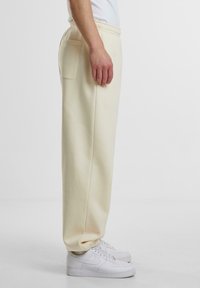 Vista lateral de una persona que lleva puesta una sudadera crema con cintura elástica y un bolsillo trasero, además de zapatillas blancas de Nike, de pie contra un fondo liso.