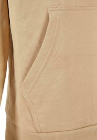 Tessuto di felpa beige con una consistenza morbida, dotato di tasca a marsupio e orlo a coste. Il design sottolinea il comfort e la vestibilità casual.