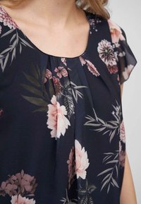 Blouse courte à manches courtes à motifs floraux en marine avec des fleurs roses et blanches. Présente des plis près de l'encolure et une texture de tissu douce.
