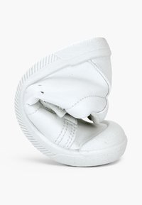 Baskets blanches flexibles pliées en deux pour montrer leur semelle et tige souples et pliables sur un fond uni.