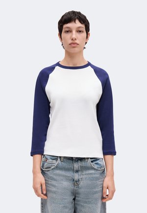 Personne aux cheveux courts et foncés portant une chemise blanche avec des manches trois-quarts bleu marine et un jean bleu, faisant face avec une expression neutre.