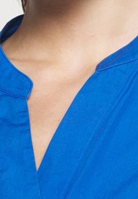 Blaues Hemd mit Kragen und V-Ausschnitt. Glattes Baumwollmaterial, das durch saubere Nähte an den Kanten besticht. Keine sichtbaren Muster.