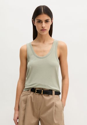 Donna con capelli lisci e scuri che indossa un top verde chiaro senza maniche, pantaloni beige a vita alta e una cintura nera con fibbia dorata.