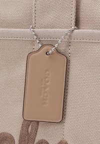 Sac en toile beige avec des détails cousus et une étiquette en cuir fauve embossée "COACH NEW YORK" attachée par une chaîne boules argentée.