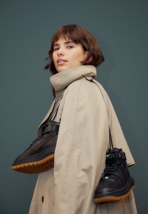 Jeune personne aux cheveux bruns courts portant un manteau beige, portant des baskets noires Nike avec semelles en gomme sur l'épaule devant un fond gris.