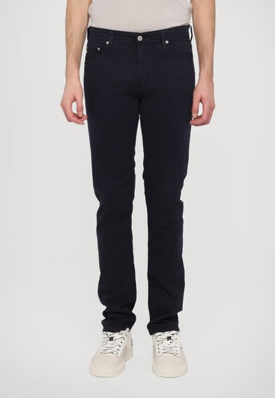 Dunkelblaue Baumwollmischung Slim-Fit-Jeans mit fünf Taschen, vorderem Knopfverschluss und minimalen Nähte-Details. Getragen mit weißen Turnschuhen.